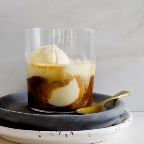 A glass cup filled with creamy vanilla ice cream, elegantly transformed into an affogato with a hot espresso pour, creates a swirling effect on a gray plate atop a white one. A small gold spoon rests nearby. The light marble surface background adds to the dessert's minimalist elegance.